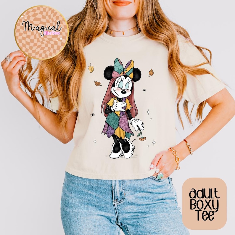 Pu&ograve; includere: T-shirt boxy adulto color crema con grafica di Minnie Mouse con abito e fiocco patchwork. Il design include foglie autunnali e piccole stelle. La maglietta ha il testo "Magical Memories" su un adesivo rotondo.