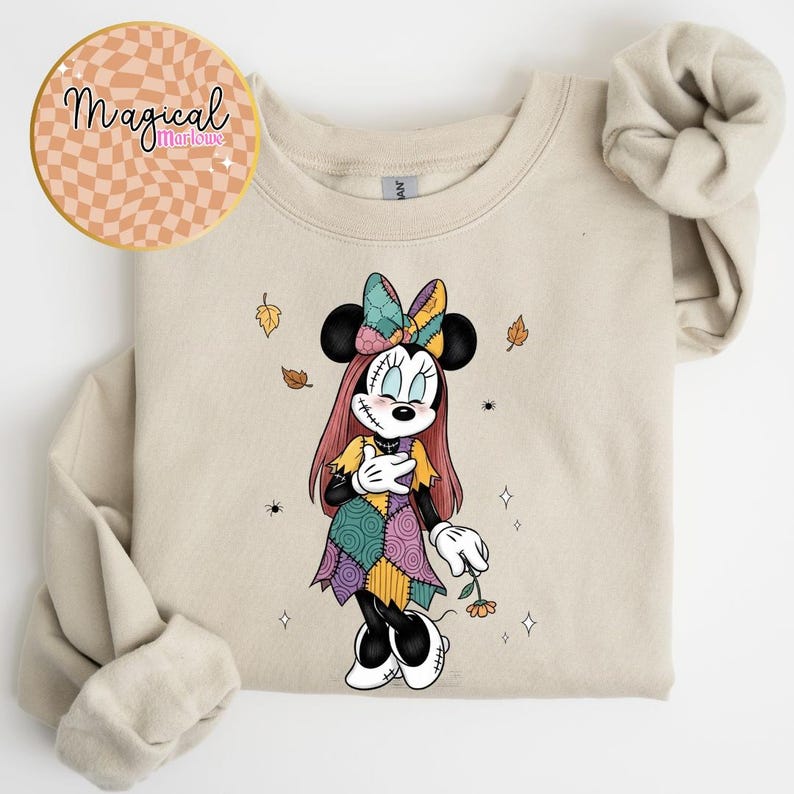Pu&ograve; includere: Felpa girocollo beige con un personaggio dei cartoni animati di Minnie Mouse in un vestito patchwork, che tiene un fiore. Il design include foglie d'autunno e il testo "Magical Marlowe" in un logo circolare.