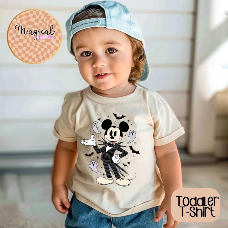Pu&ograve; includere: Un bambino piccolo indossa una maglietta beige con una grafica di Mickey Mouse a tema Halloween. Il design presenta Mickey in un abito gessato con pipistrelli e fantasmi. La maglietta ha anche la scritta "Toddler T-Shirt". Il bambino indossa un berretto da baseball azzurro.