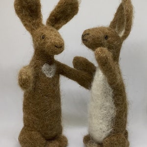 May include: Two brown felt bunny rabbits standing side by side. One bunny has a white heart on its chest.