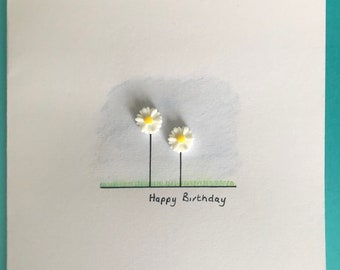 May include: A white card with a simple drawing of two white daisies with yellow centers and green grass. The text "Happy Birthday" is written in black ink below the daisies.