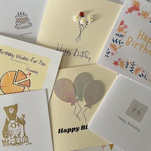 May include: A selection of birthday cards with diverse designs. The cards include illustrations of a cake, balloons, a bear, and flowers. Handwritten messages such as "Happy Birthday" and "Birthday Wishes For You" are visible.
