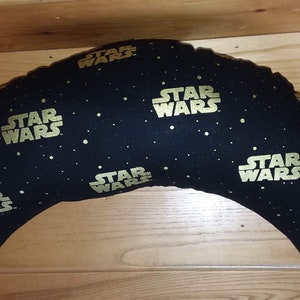 May include: A crescent-shaped pillow with a black fabric and a gold "STAR WARS" print. The pillow is covered in small gold dots.