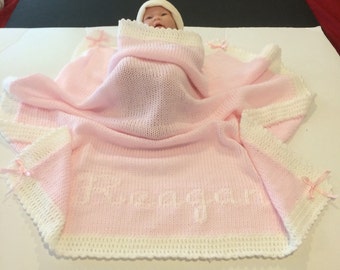 May include: A baby wrapped in a pink knitted blanket with white trim and the name "Reagan" embroidered on it. The blanket has a scalloped edge and is adorned with small pink bows. A white hat covers the baby's head.