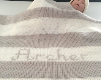May include: A knitted baby blanket with alternating stripes of white and gray. The name "Archer" is subtly embroidered in the center. A baby doll wearing a white hat is partially visible, wrapped in the blanket.