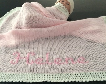 May include: A baby wrapped in a pink knitted blanket with the name "Helena" embroidered in pink. The blanket has a white crocheted border and a matching white hat. The baby is nestled in the blanket.