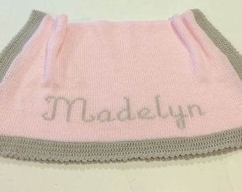 May include: A pink knitted baby blanket with a gray crocheted border. The name "Madelyn" is embroidered in gray across the center of the blanket. The blanket is folded, showing the name and border.