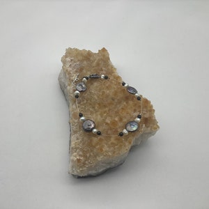 May include: A delicate bracelet featuring iridescent, flat, round pearls and small black beads strung on a thin, clear wire. The bracelet is displayed on a rough, light-colored stone, showcasing its elegant design.