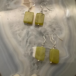 May include: Four pairs of rectangular, pale green earrings with silver hooks. The earrings are made of a translucent, light green stone. The earrings are displayed on a light grey and white surface.