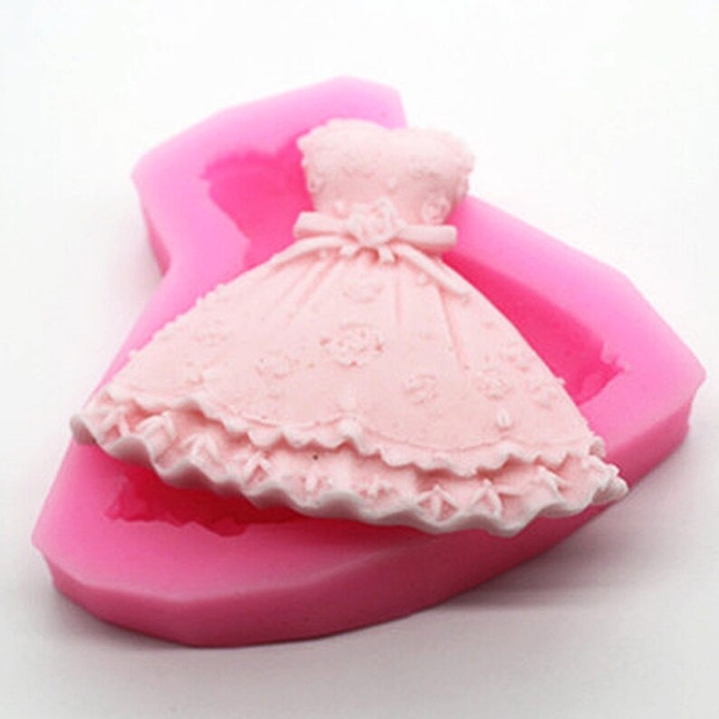 May include: Pink silicone mould shaped like a dress with a lace trim and floral design. The mould is designed for making cake decorations or other edible treats.