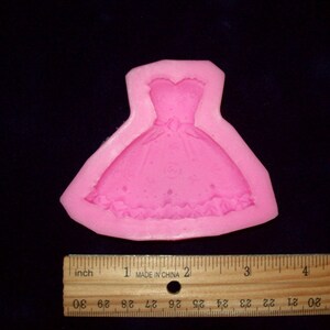 May include: Pink silicone mould shaped like a dress with a ruffled bottom. The mould is approximately 5 cm wide and 6.5 cm tall.