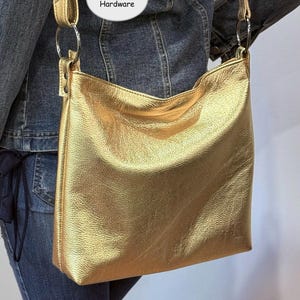 Puede incluir: Un bolso de hombro dorado con una correa larga y herrajes de metal pavonado. El bolso tiene forma rectangular y un aspecto suave y texturizado. El texto "Gunmetal Hardware" es visible en la imagen.