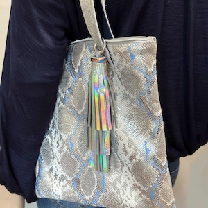 May include: A silver tote bag with a snakeskin pattern in gray, white, and blue. The bag has a silver strap and a decorative tassel with iridescent fringe. The bag is being carried over the shoulder.