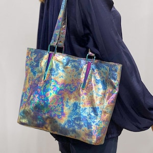 May include: A rectangular tote bag with a metallic, iridescent finish featuring swirling patterns of blue, gold, and purple. The bag includes two shoulder straps attached with shiny, dark-toned hardware that matches the bag's reflective surface.