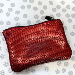 May include: A small, rectangular red sequin pouch with a black zip and pull. The pouch is on a white background with a pattern of evenly spaced grey polka dots. The sequins give the pouch a textured, shimmering appearance.