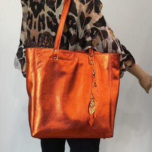 May include: A metallic orange tote bag with two shoulder straps and a decorative leaf-shaped charm hanging from one of the strap attachments. The bag features a smooth, reflective finish and is constructed with visible stitching along the sides.