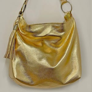 Puede incluir: Un bolso de hombro dorado con una sola correa y una borla. El bolso tiene un dise&ntilde;o suave y holgado y un acabado brillante. La correa est&aacute; sujeta al bolso con un anillo plateado.