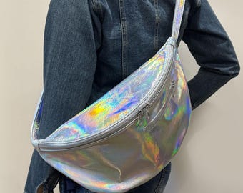 May include: A large crescent-shaped crossbody bag featuring a reflective, iridescent holographic finish that shifts between silver, blue, and pink tones. The bag includes a wide adjustable strap and a front zipper closure with two silver metal pulls.