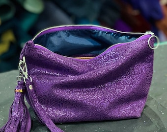 May include: A purple clutch bag featuring a subtle embossed floral pattern and a shimmering finish. The bag includes a silver-toned zipper closure, a matching purple tassel attached to a metal clip, and a dark blue interior lining.