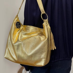 Puede incluir: Un bolso de hombro de cuero texturizado de color dorado con una correa larga y una borla. El bolso tiene un dise&ntilde;o grande y abierto y un detalle de anillo circular donde se sujeta la correa. Una peque&ntilde;a etiqueta cuelga del bolso.