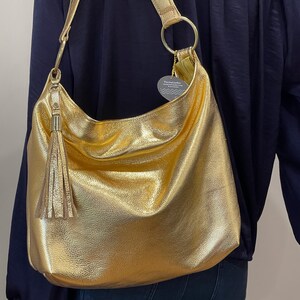 Puede incluir: Un bolso de hombro dorado con una correa larga y una borla. El bolso tiene una forma grande y redondeada y un brillo met&aacute;lico. La correa est&aacute; sujeta al bolso con anillos dorados.