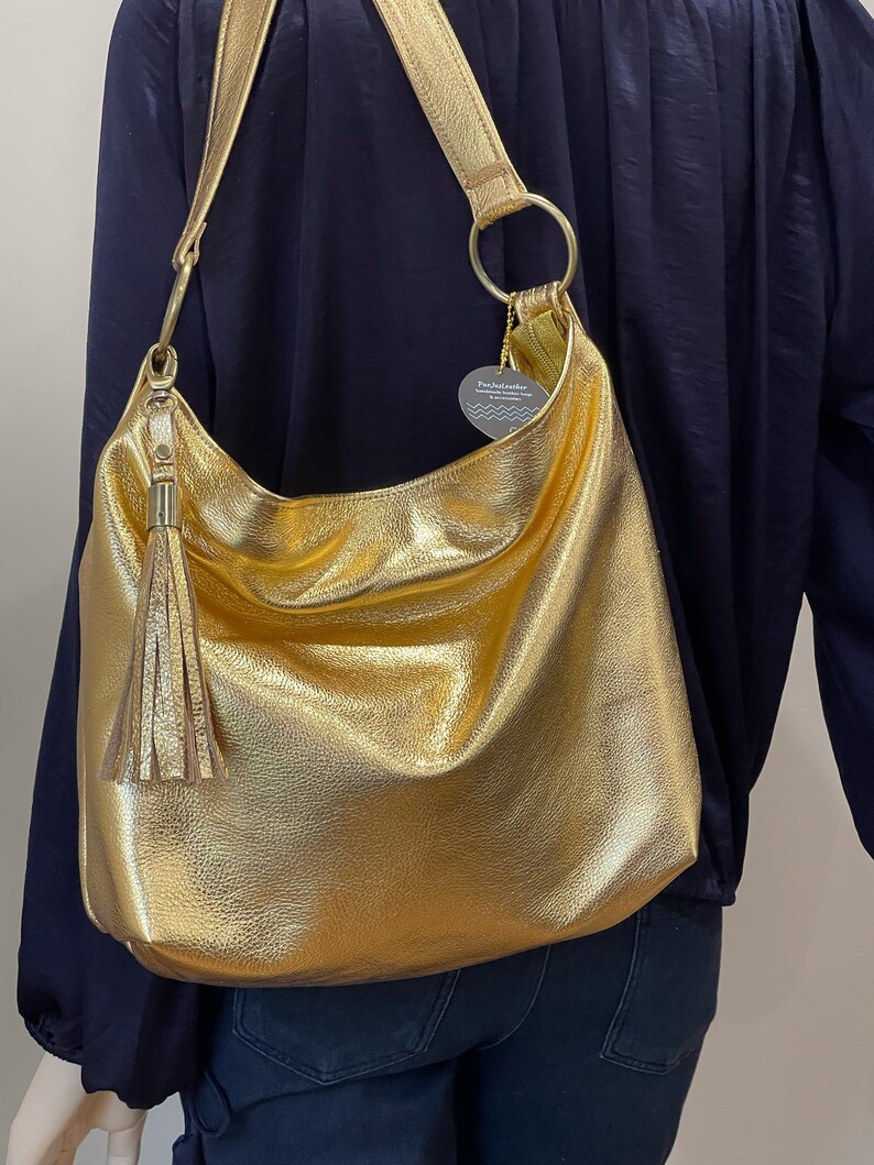 Puede incluir: Un bolso de hombro dorado con una correa larga y una borla. El bolso tiene una forma grande y redondeada y un brillo met&aacute;lico. La correa est&aacute; sujeta al bolso con anillos dorados.