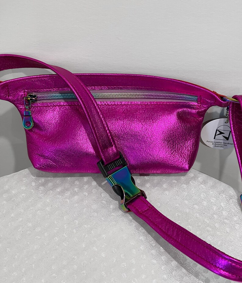 Puede incluir: Una ri&ntilde;onera de cuero iridiscente rosa brillante con un bolsillo con cremallera y una correa ajustable con una hebilla plateada. La bolsa tiene una peque&ntilde;a etiqueta que dice "Pure Leather Handbags & Accessories Australia".
