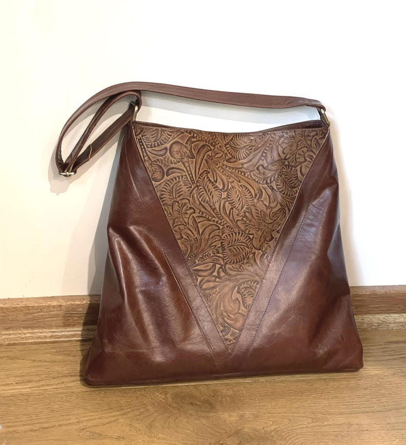 May include: A brown leather shoulder bag featuring a large triangular panel with an intricate embossed floral pattern on the front. The bag has a smooth finish on the lower sections and sides, complemented by a single adjustable shoulder strap.
