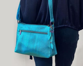 May include: A teal crossbody bag with a metallic finish. It has a zip-up main compartment and an adjustable strap. A decorative feather charm hangs from the side. The bag is made of a textured material.
