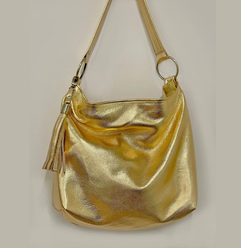 Puede incluir: Un bolso de hombro dorado con una sola correa y una borla. El bolso tiene un dise&ntilde;o suave y holgado y un acabado brillante. La correa est&aacute; sujeta al bolso con un anillo plateado.