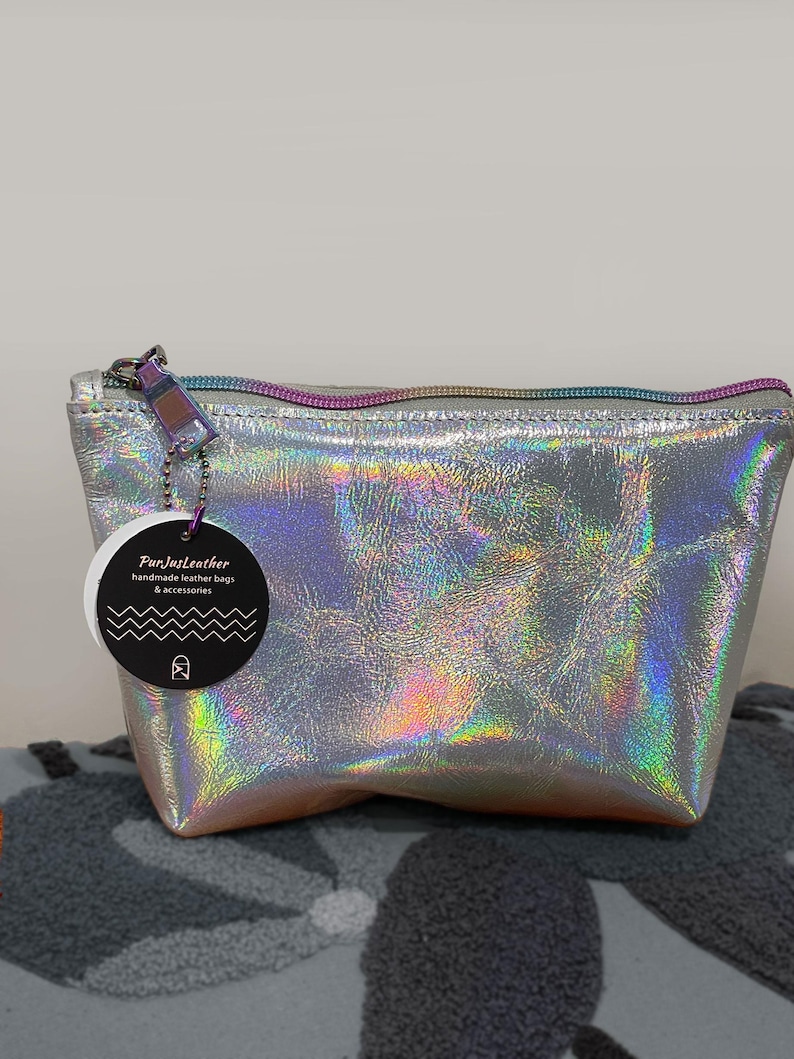 May include: A silver holographic leather pouch with a rainbow zipper and a tag that reads "FunJustLeather handmade leather bags & accessories". The pouch is sitting on a gray patterned surface.