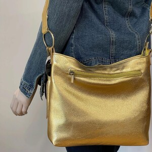 Puede incluir: Un bolso bandolera dorado con cierre de cremallera y una correa larga. El bolso tiene una superficie texturizada y un detalle de anillo circular donde se conecta la correa. El bolso lo lleva una persona con una chaqueta vaquera.
