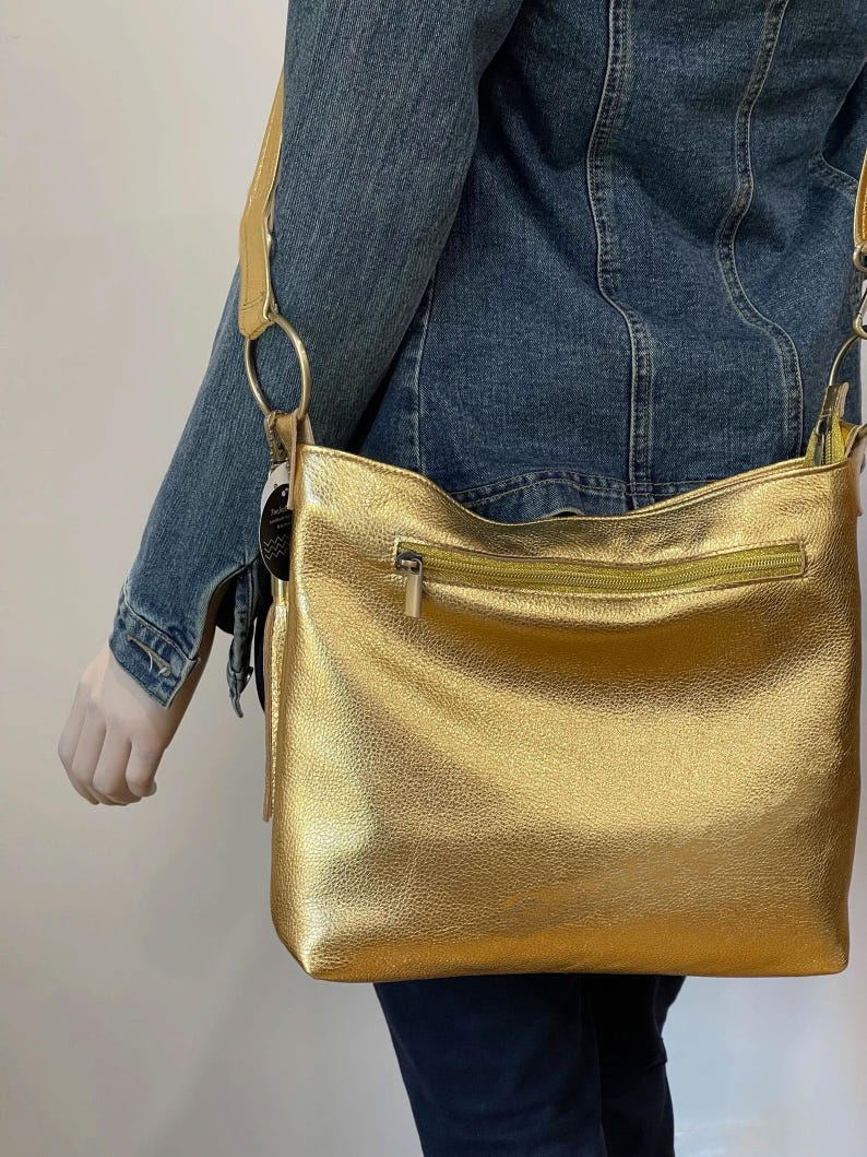 Puede incluir: Un bolso bandolera dorado con cierre de cremallera y una correa larga. El bolso tiene una superficie texturizada y un detalle de anillo circular donde se conecta la correa. El bolso lo lleva una persona con una chaqueta vaquera.