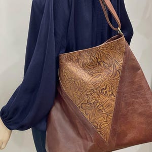 May include: A brown shoulder bag featuring a large triangular center panel with an intricate embossed floral pattern. The bag has smooth brown side panels and an adjustable brown strap with metallic hardware, worn over a dark blue long-sleeve top.