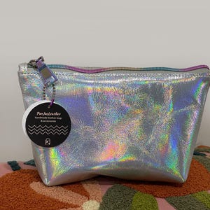 May include: A silver holographic leather bag with a rainbow zipper and a tag that reads "FunJustLeather handmade leather bags & accessories". The bag is sitting on a pink and orange patterned surface.