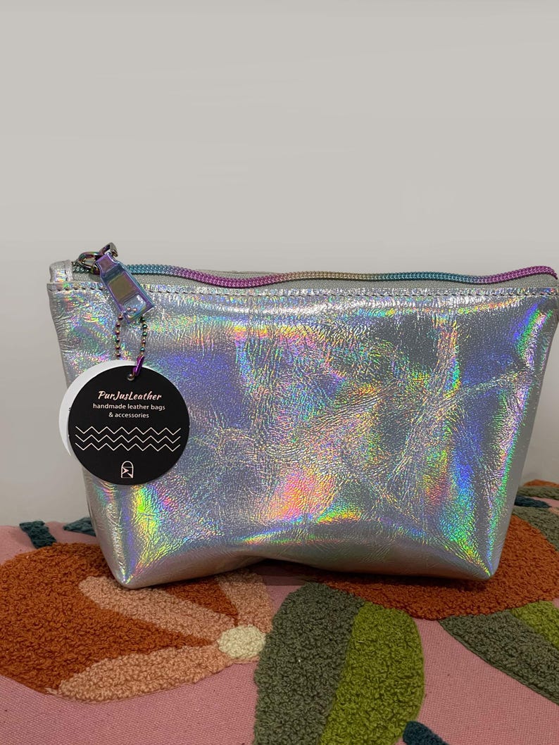 May include: A silver holographic leather bag with a rainbow zipper and a tag that reads "FunJustLeather handmade leather bags & accessories". The bag is sitting on a pink and orange patterned surface.