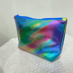 Puede incluir: Una bolsa de maquillaje hologr&aacute;fica azul con un patr&oacute;n de remolino arco&iacute;ris. La bolsa tiene una cremallera.