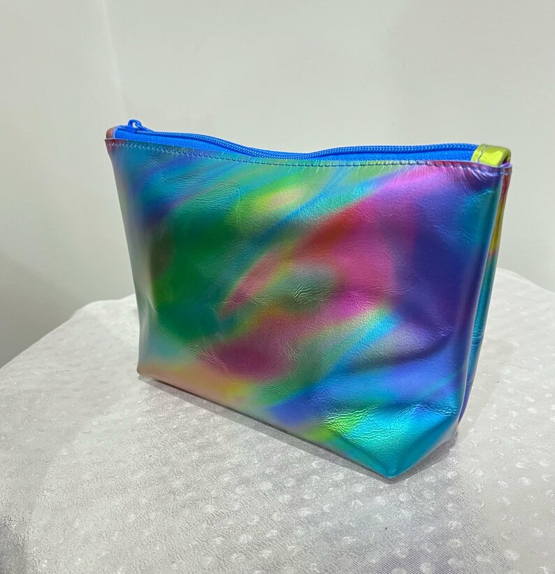 Puede incluir: Una bolsa de maquillaje hologr&aacute;fica azul con un patr&oacute;n de remolino arco&iacute;ris. La bolsa tiene una cremallera.