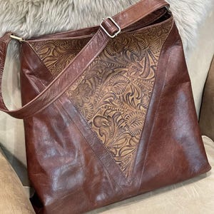 May include: A brown leather shoulder bag featuring a large triangular center panel with an intricate embossed floral pattern. The bag includes a matching brown leather adjustable strap with a silver-toned metal buckle and smooth leather side panels.
