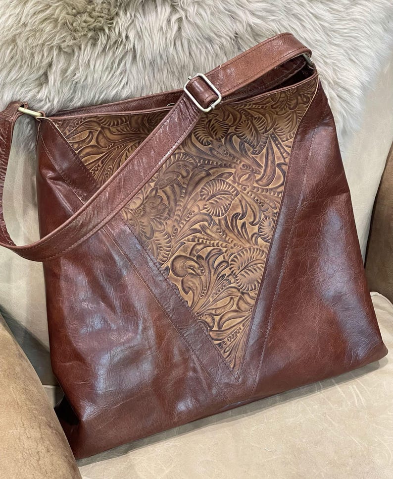 May include: A brown leather shoulder bag featuring a large triangular center panel with an intricate embossed floral pattern. The bag includes a matching brown leather adjustable strap with a silver-toned metal buckle and smooth leather side panels.