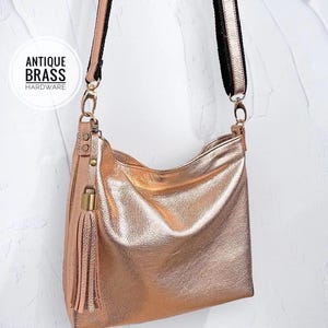 May include: A rose gold leather shoulder bag with a tassel and antique brass hardware. The bag has a black and rose gold adjustable strap.