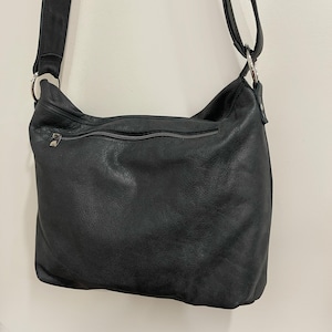 May include: Black leather shoulder bag with a long adjustable strap. The bag has a zippered top closure and a zippered pocket on the back. The leather appears to be soft and has a slightly textured finish. The bag is hanging against a plain white wall.