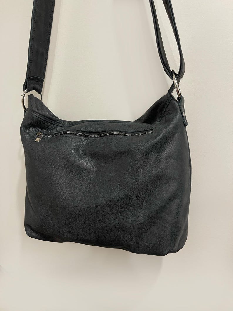 May include: Black leather shoulder bag with a long adjustable strap. The bag has a zippered top closure and a zippered pocket on the back. The leather appears to be soft and has a slightly textured finish. The bag is hanging against a plain white wall.