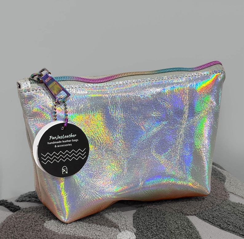 May include: Silver holographic leather bag with a rainbow zipper and a tag that reads "FunJustLeather handmade leather bags & accessories". The bag has a rectangular shape with a slight trapezoid design. The zipper pull is a small rectangle with a rainbow finish.