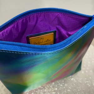 Puede incluir: Una peque&ntilde;a bolsa iridiscente azul, verde y rosa con cierre de cremallera. La bolsa tiene un forro morado y una etiqueta de cuero marr&oacute;n con el texto "Paravel Leather" en letras doradas.
