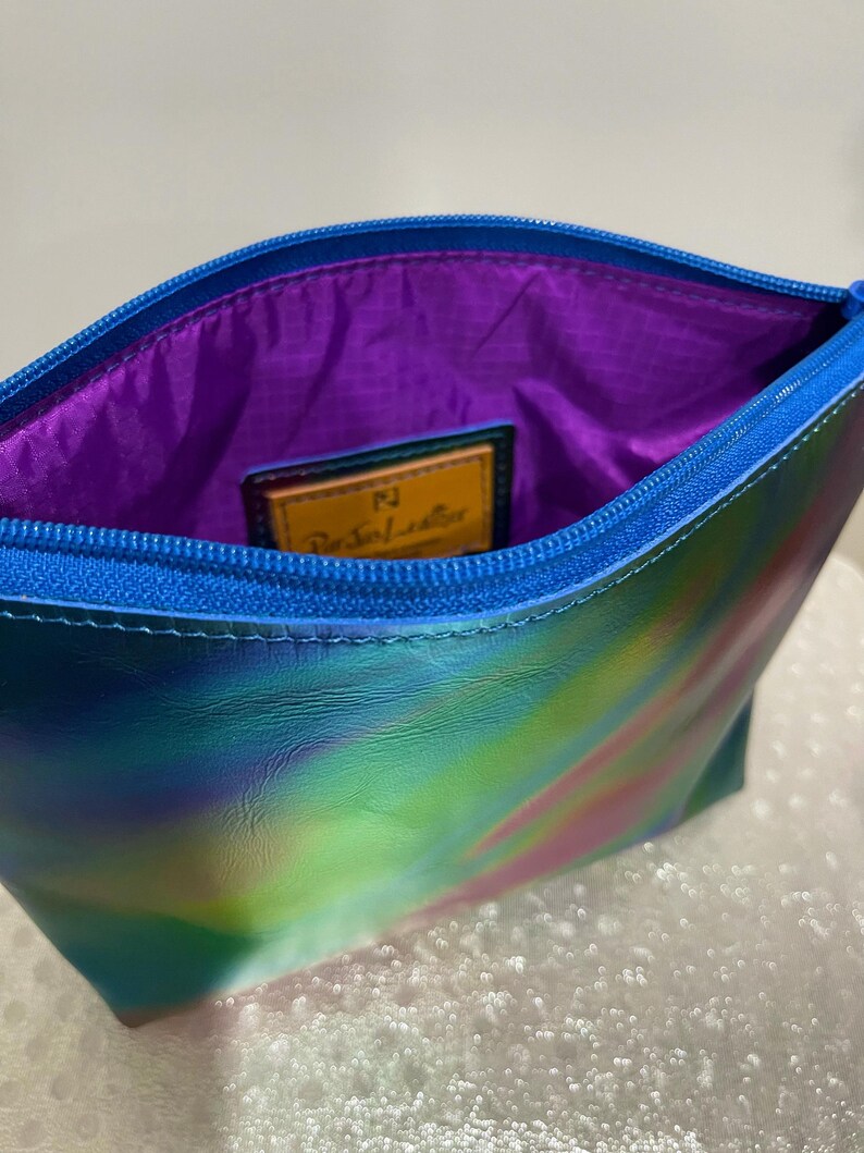Puede incluir: Una peque&ntilde;a bolsa iridiscente azul, verde y rosa con cierre de cremallera. La bolsa tiene un forro morado y una etiqueta de cuero marr&oacute;n con el texto "Paravel Leather" en letras doradas.