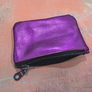 K&ouml;nnte beinhalten: Ein rechteckiges Rei&szlig;verschlussetui aus metallisch violettem Leder mit sichtbarer Narbenstruktur. Das Etui verf&uuml;gt &uuml;ber ein schwarzes Innenfutter und einen schwarzen Rei&szlig;verschluss mit einem metallisch schillernden Zipper am Schieber.