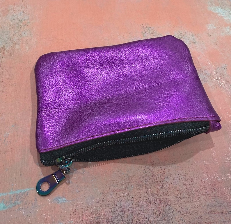 K&ouml;nnte beinhalten: Ein rechteckiges Rei&szlig;verschlussetui aus metallisch violettem Leder mit sichtbarer Narbenstruktur. Das Etui verf&uuml;gt &uuml;ber ein schwarzes Innenfutter und einen schwarzen Rei&szlig;verschluss mit einem metallisch schillernden Zipper am Schieber.