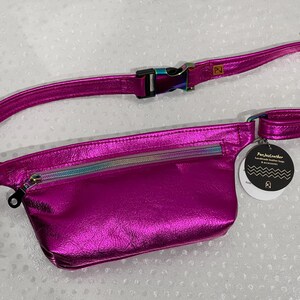 Puede incluir: Una ri&ntilde;onera de cuero met&aacute;lico rosa brillante con un bolsillo con cremallera y una cremallera arco&iacute;ris iridiscente. La ri&ntilde;onera tiene un cintur&oacute;n negro ajustable con una hebilla plateada. La etiqueta adjunta a la ri&ntilde;onera dice "Pur.JustLeather handmade leather bags & accessories".