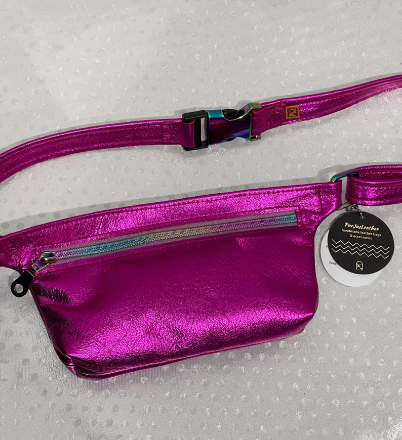 Puede incluir: Una ri&ntilde;onera de cuero met&aacute;lico rosa brillante con un bolsillo con cremallera y una cremallera arco&iacute;ris iridiscente. La ri&ntilde;onera tiene un cintur&oacute;n negro ajustable con una hebilla plateada. La etiqueta adjunta a la ri&ntilde;onera dice "Pur.JustLeather handmade leather bags & accessories".
